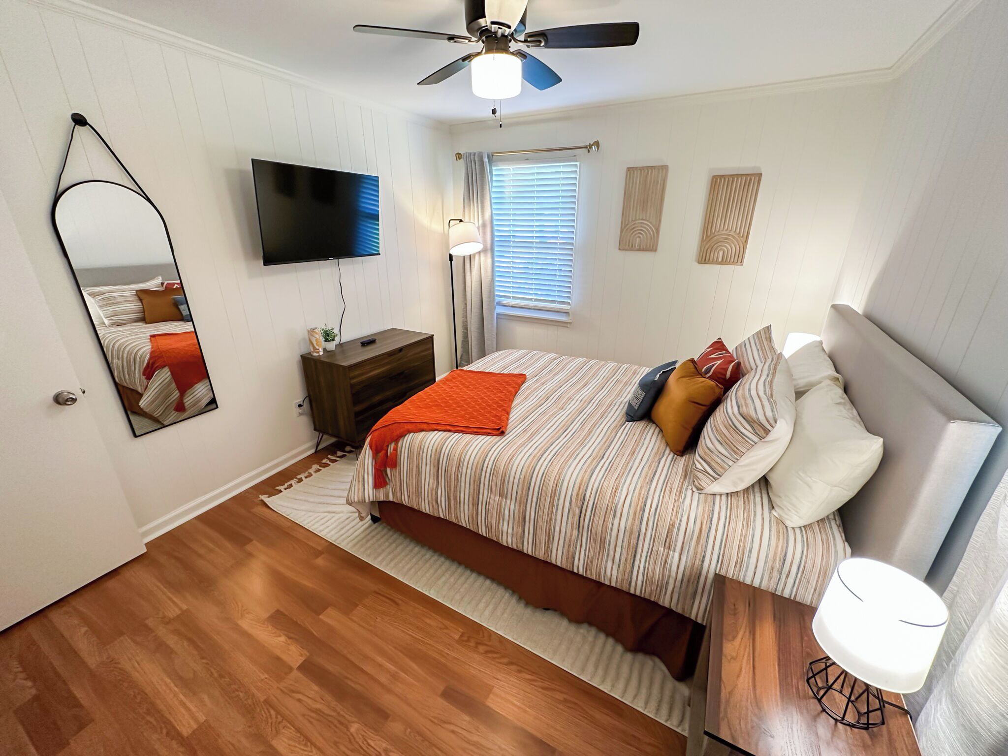 A bedroom with a bed, a TV, a ceiling fan, a nightstand, a mirror, and a window with blinds.