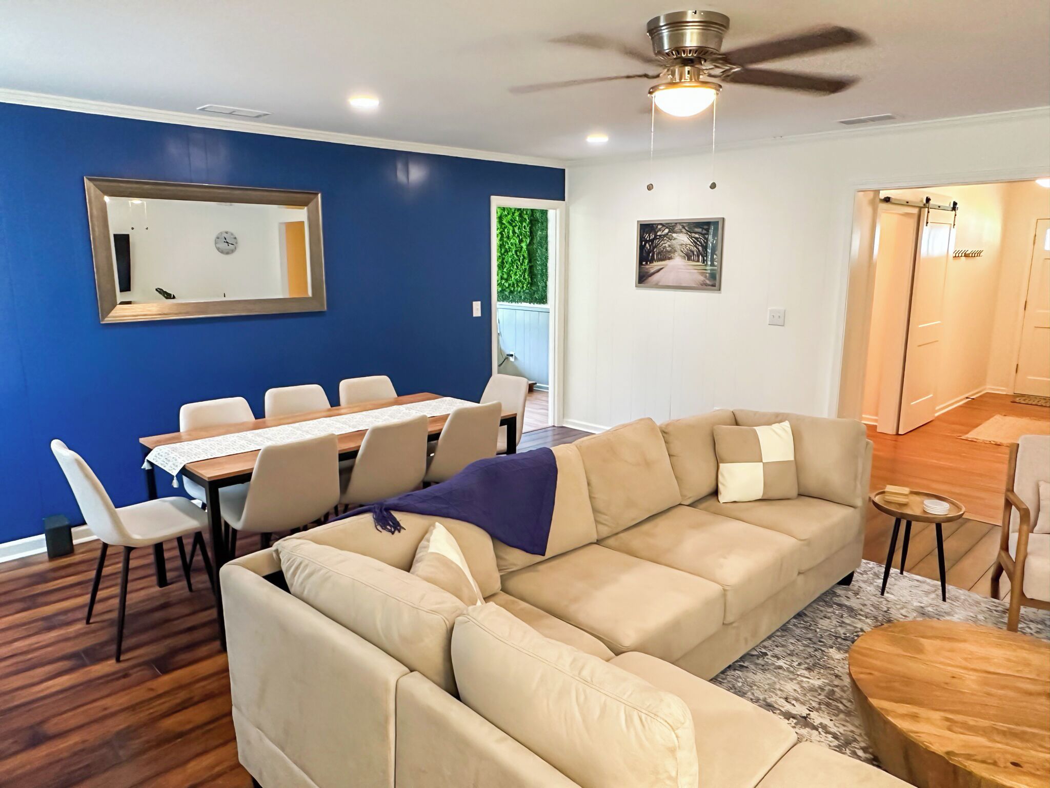 Una sala de estar con un sofá seccional color beige, una mesa de comedor con sillas, un ventilador de techo y una pequeña mesita de madera.