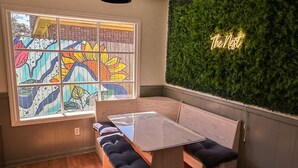 Una habitación moderna con un banco de madera, una mesa de cristal, un ventanal de vitral colorido y una pared verde con un letrero de neón.