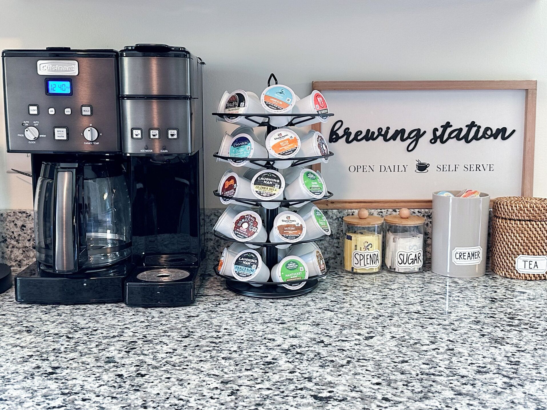 Une machine à café, un présentoir pour capsules de café et des bocaux étiquetés contenant du sucre, de la crème et du thé, le tout posé sur un plan de travail de cuisine.