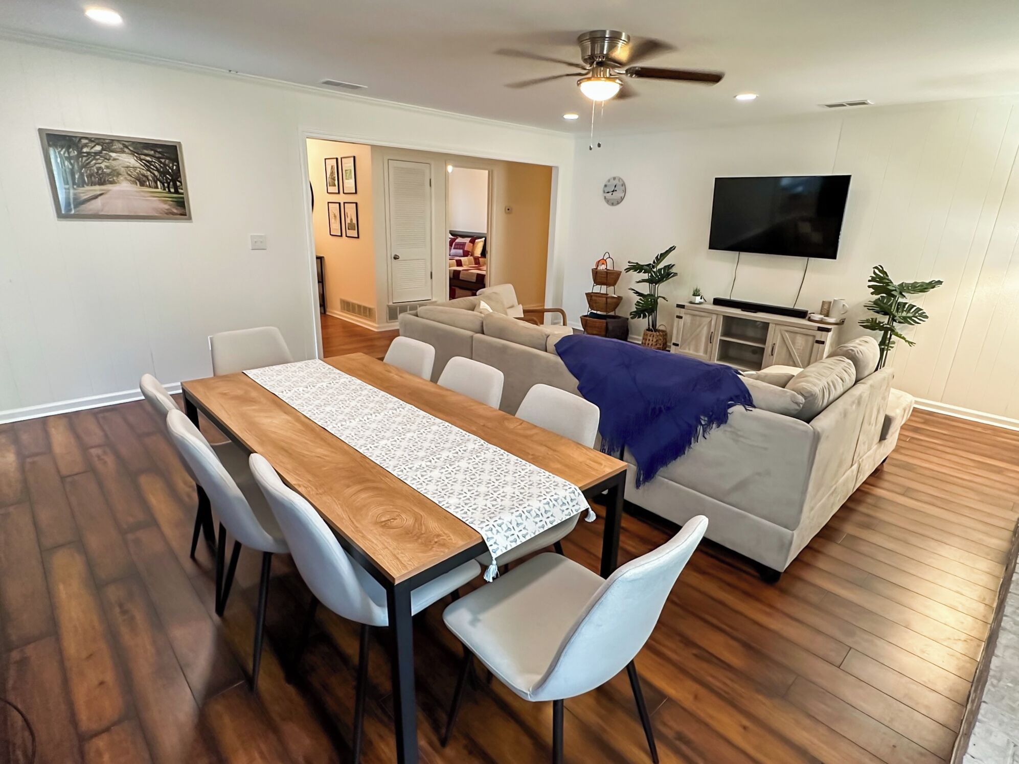 A dining area with a wooden table and white chairs, a living room with a sofa and a TV, and a ceiling fan.