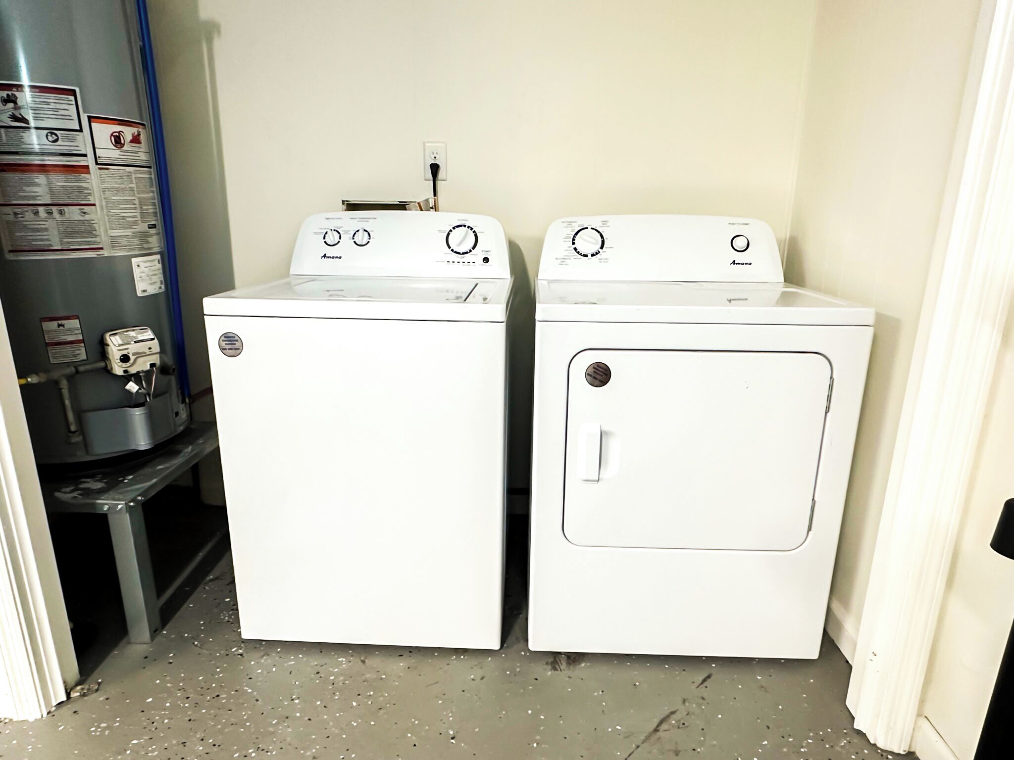 Two white front-loading washing machines in a laundry room with a water heater and a metal shelf.