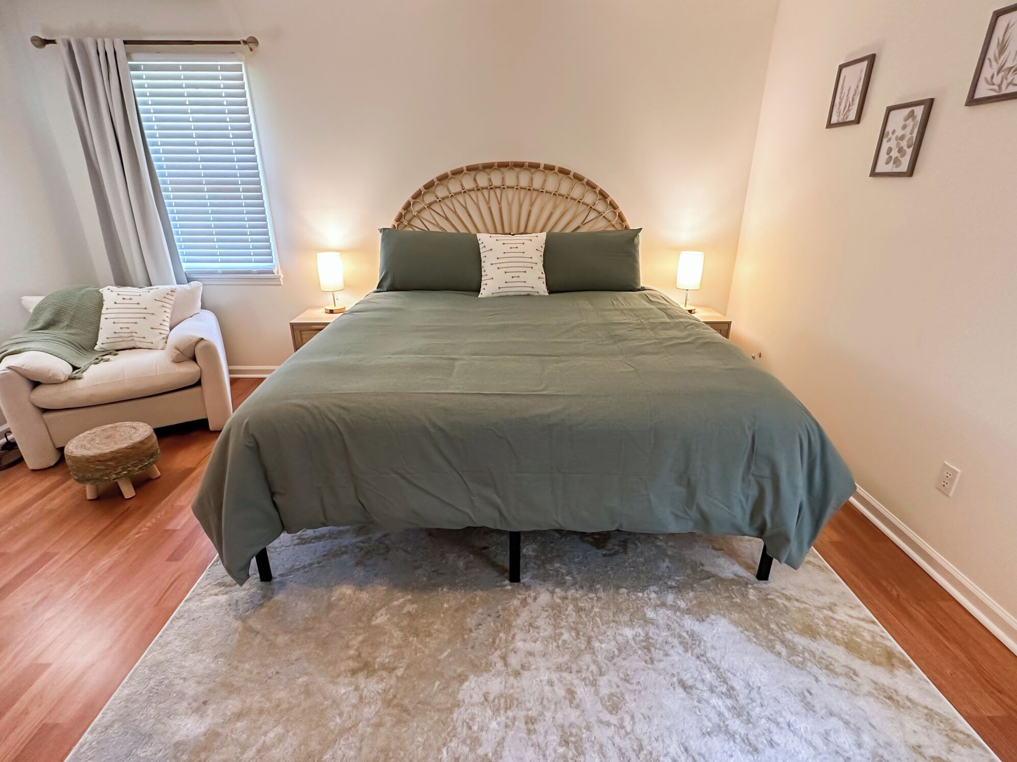 A bedroom with a large bed, two bedside lamps, a wicker headboard, a small round ottoman, and a window with blinds.