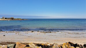 A coastal beach with rocky foreground, clear blue water, and a distant shoreline with buildings. - Lovely home in Saint-Vaast-la-Hougue with WiFi (Saint-Vaast-la-Hougue)