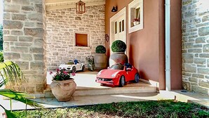 Een stenen gebouw met een rode sportauto ervoor. Planten in potten en een kleine witte auto op de achtergrond.