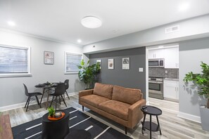 Una sala moderna con un sofá marrón, sillas negras, una mesa de comedor y una mesita auxiliar negra pequeña. Se ve un área de cocina al fondo con armarios blancos y electrodomésticos de acero inoxidable.