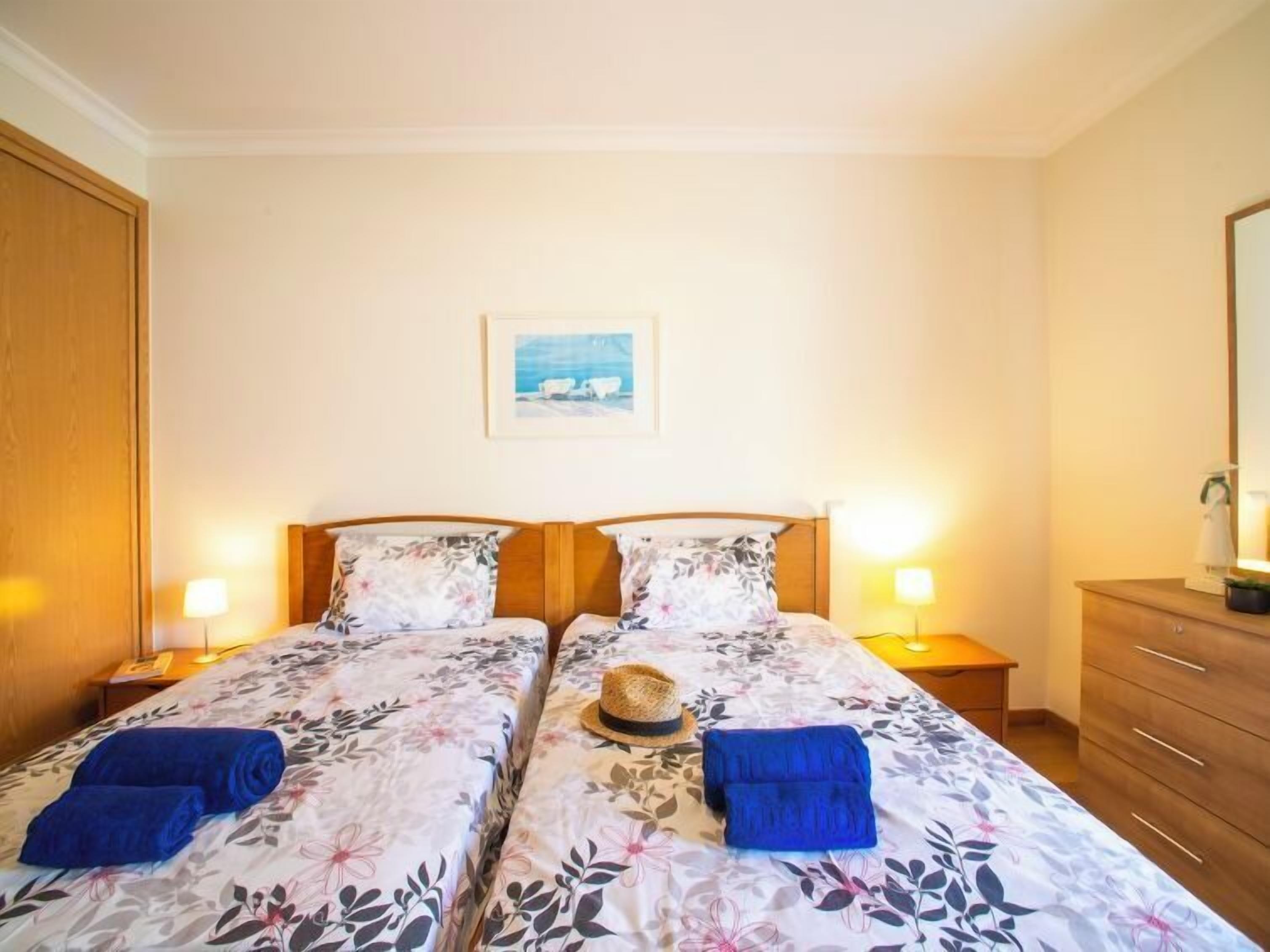 A double room with two beds, each with a floral bedspread, a wooden headboard, and bedside lamps. There is a framed picture on the wall above the beds, and a wooden dresser with a mirror to the right.