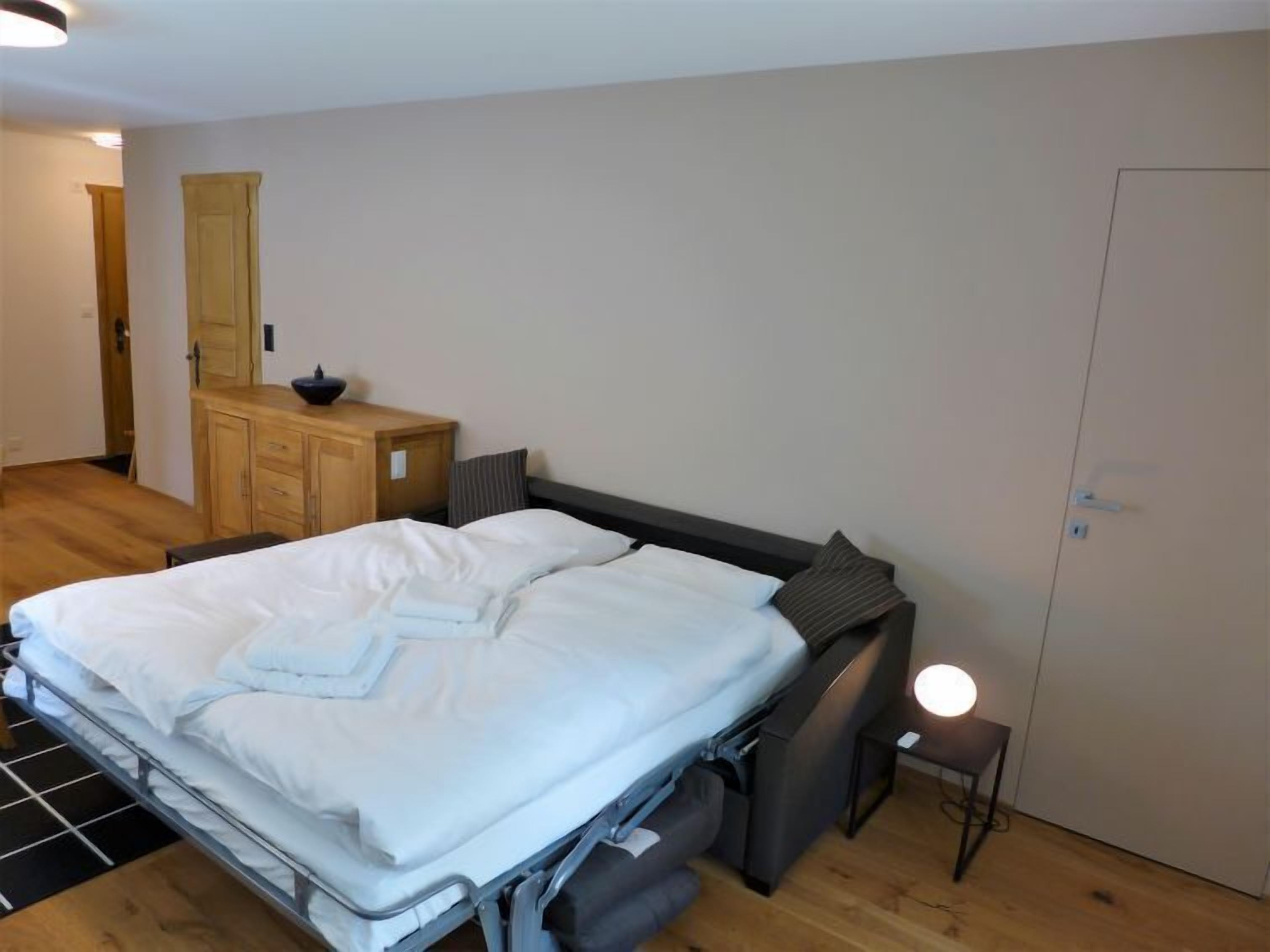 A bedroom with a bed, a wooden dresser, a small bedside table with a lamp, and a door.