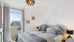 Ein Schlafzimmer mit einem großen Bett, einem Fenster mit Vorhängen, einer Deckenleuchte und einem an der Wand hängenden Bild.