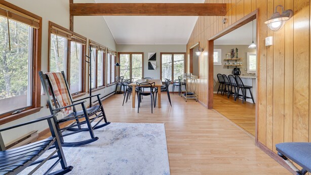 Una habitación amplia con suelos de madera, una mesa de comedor con sillas, una mecedora y grandes ventanas que ofrecen vistas al exterior.