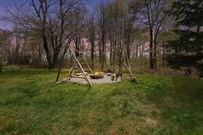 Un espace extérieur aménagé dans un style rustique, avec un foyer, entouré d’arbres et d’un champ d’herbe.
