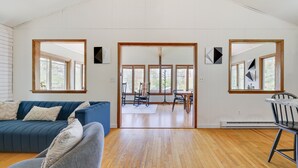 Amplia sala de estar con un sofá azul, un sillón gris y una zona de comedor con mesa y sillas. Grandes ventanas ofrecen luz natural y vistas al exterior.