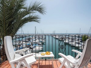 Un balcon avec des chaises blanches, une petite table sur laquelle se trouve un objet jaune, et une vue sur une marina avec des bateaux.