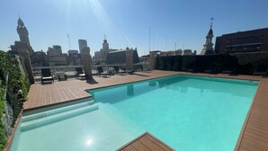A rooftop swimming pool with wooden deck and lounge chairs, overlooking a cityscape with various buildings and a clear sky.