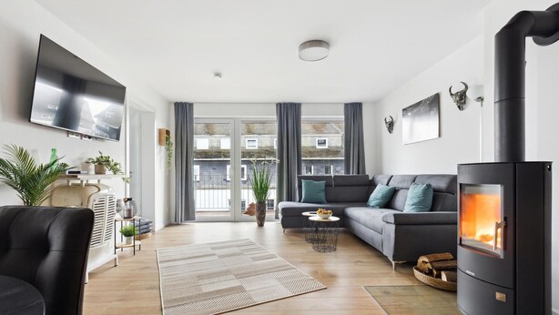 A modern living room with a sofa, a TV, a fireplace, and a view of buildings through large windows. - GebirgsResort Winterberg (Winterberg)