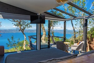 A modern bedroom with a large bed, a TV, and a view of a lake and mountains through large windows.
