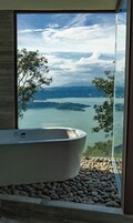 A modern bathroom with a freestanding bathtub, a stone-tiled floor, and a large window offering a view of a lake and distant mountains.