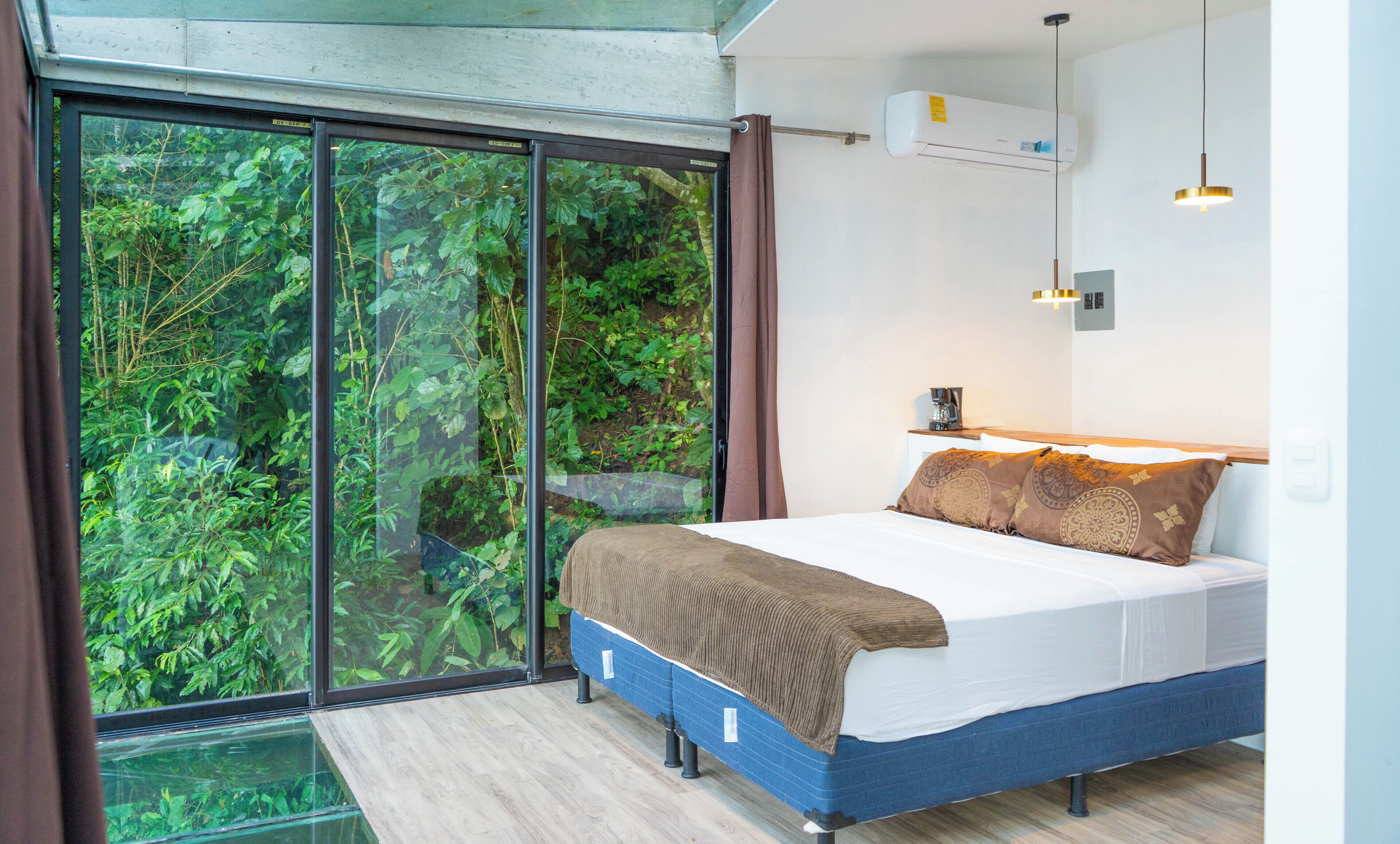 A bedroom with a large sliding glass door leading to a lush green forest view, a bed with a brown blanket, a coffee maker on a bedside table, and an air conditioning unit on the wall.