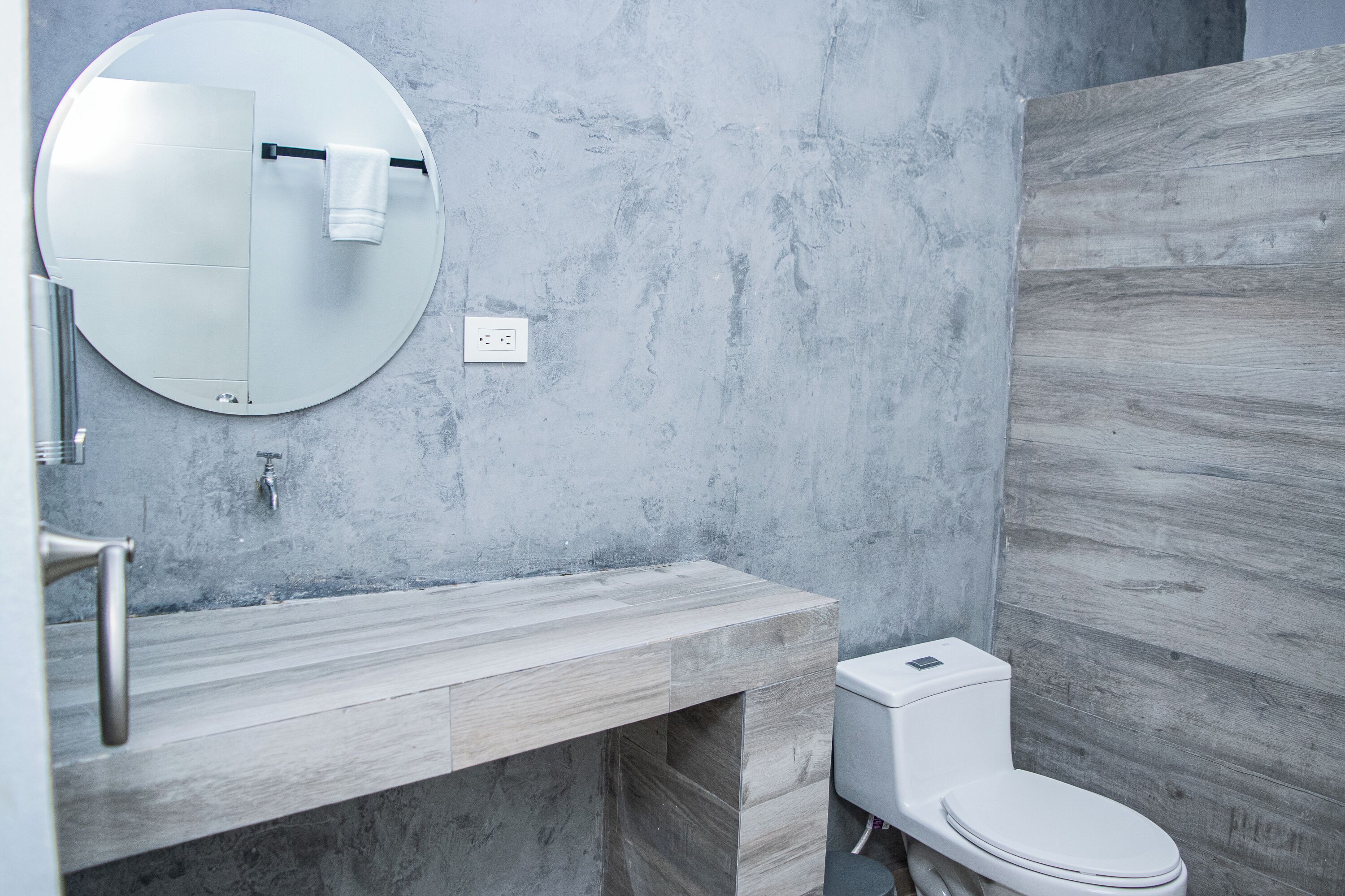 A modern bathroom with a round mirror, a wooden vanity, a toilet, and a wall-mounted towel rack.