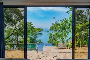 A view from a room with a large window overlooking a pool and lush greenery.