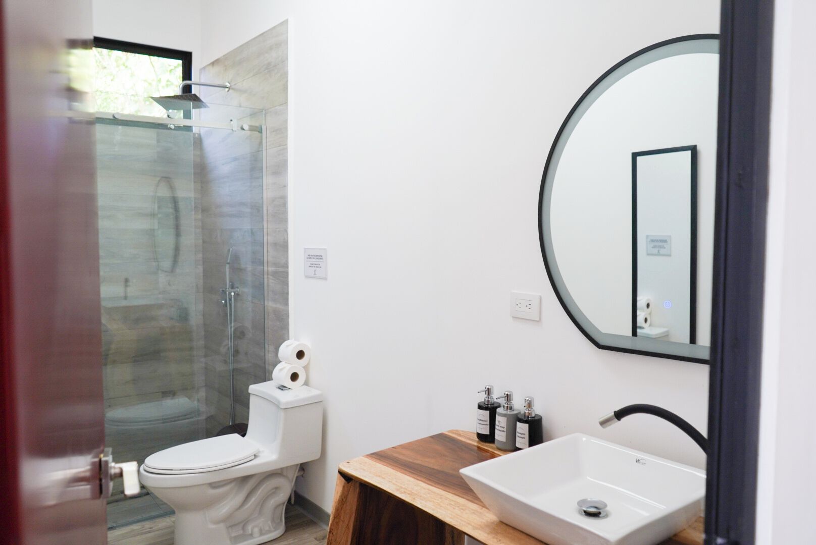 Ein modernes Badezimmer mit Duschkabine aus Glas, einer weißen Toilette, einem hölzernen Waschtisch und einem runden Spiegel.