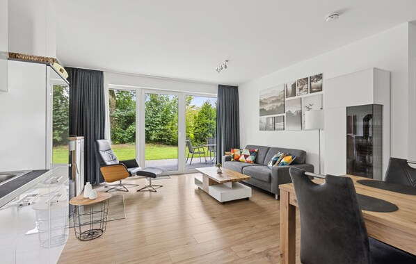 Ein modernes Wohnzimmer mit Sofa, Couchtisch und Sesseln. Ein großes Fenster mit Vorhängen, ein Kamin und ein Essbereich mit Tisch und Stühlen sind ebenfalls vorhanden.