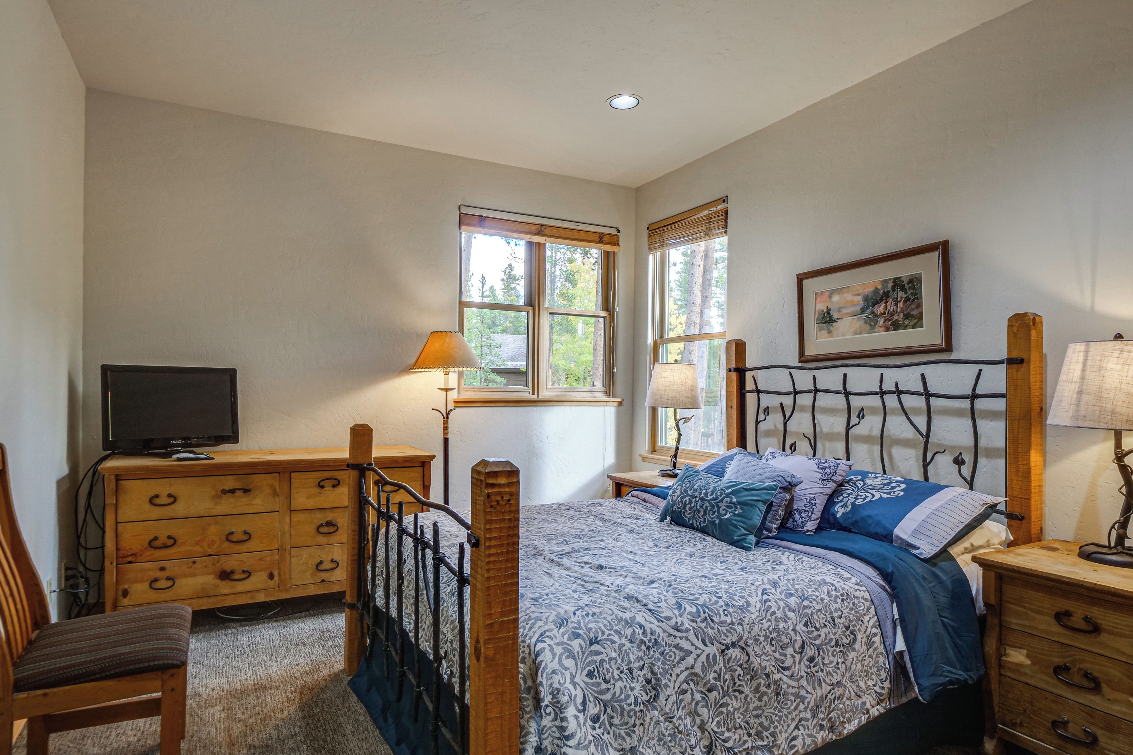 Une chambre à coucher comprenant un lit, une commode en bois, une chaise, un téléviseur et deux fenêtres.
