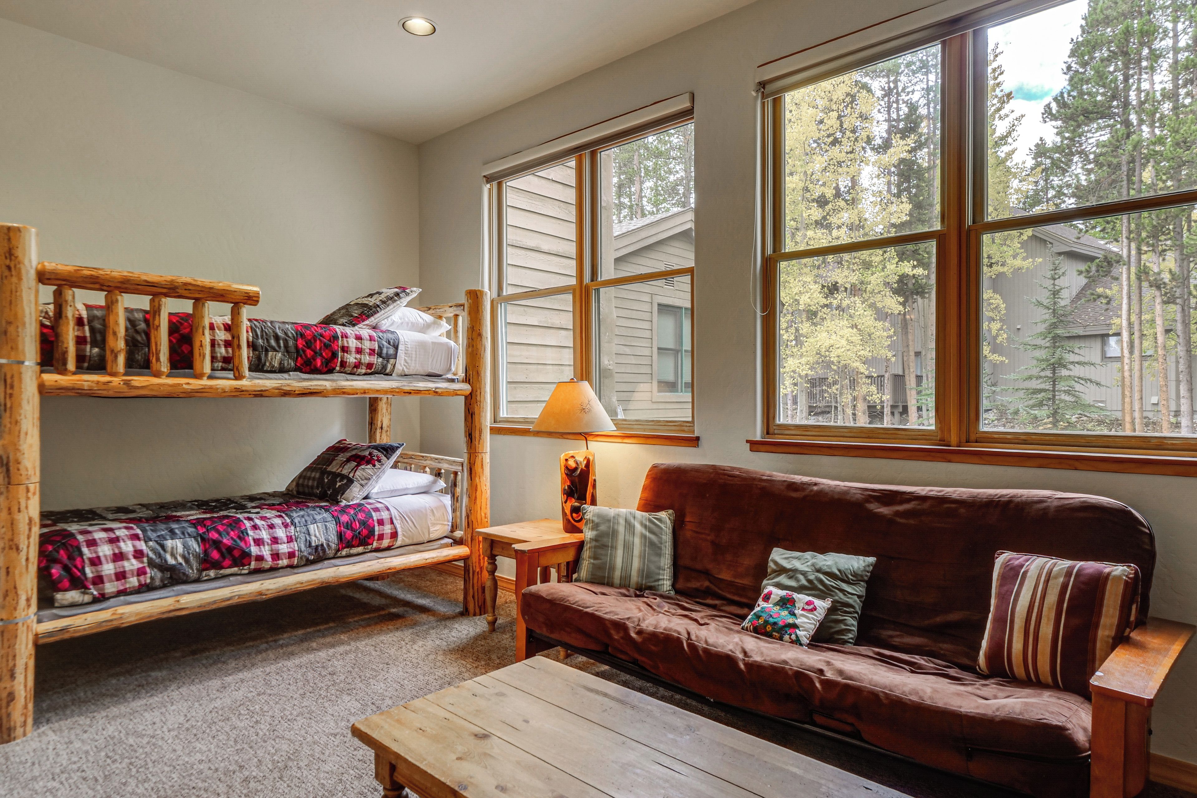 A room with a bunk bed, a couch, and a table. There are large windows providing a view of trees and a building outside.