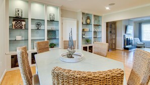 Un comedor con una mesa blanca, sillas de mimbre y repisas de vidrio con adornos.