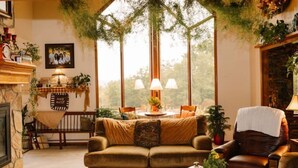 Una sala de estar acogedora con chimenea, un sofá, una mesa de comedor con sillas y vistas a árboles a través de amplias ventanas.