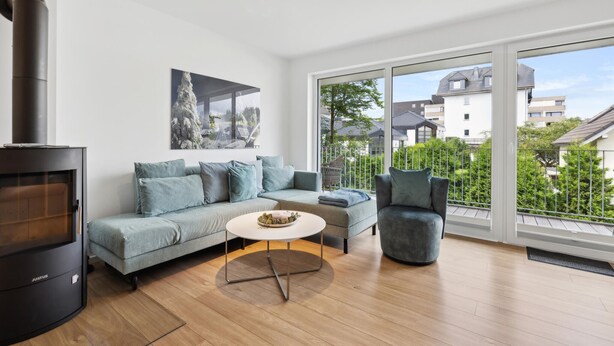 A modern living room with a Justus wood-burning stove, a grey sofa, a round coffee table, and a view of a residential area through large windows.