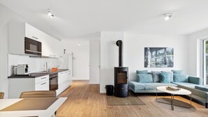 A modern kitchen with a built-in oven, microwave, and coffee machine. There is a black wood-burning stove in the living area, a sofa with blue cushions, and a painting on the wall.