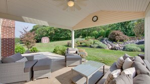 A covered patio area with wicker furniture, a glass table, and a view of a landscaped garden. - Game Room & Covered Patio: Hummelstown Hideaway (Hummelstown)