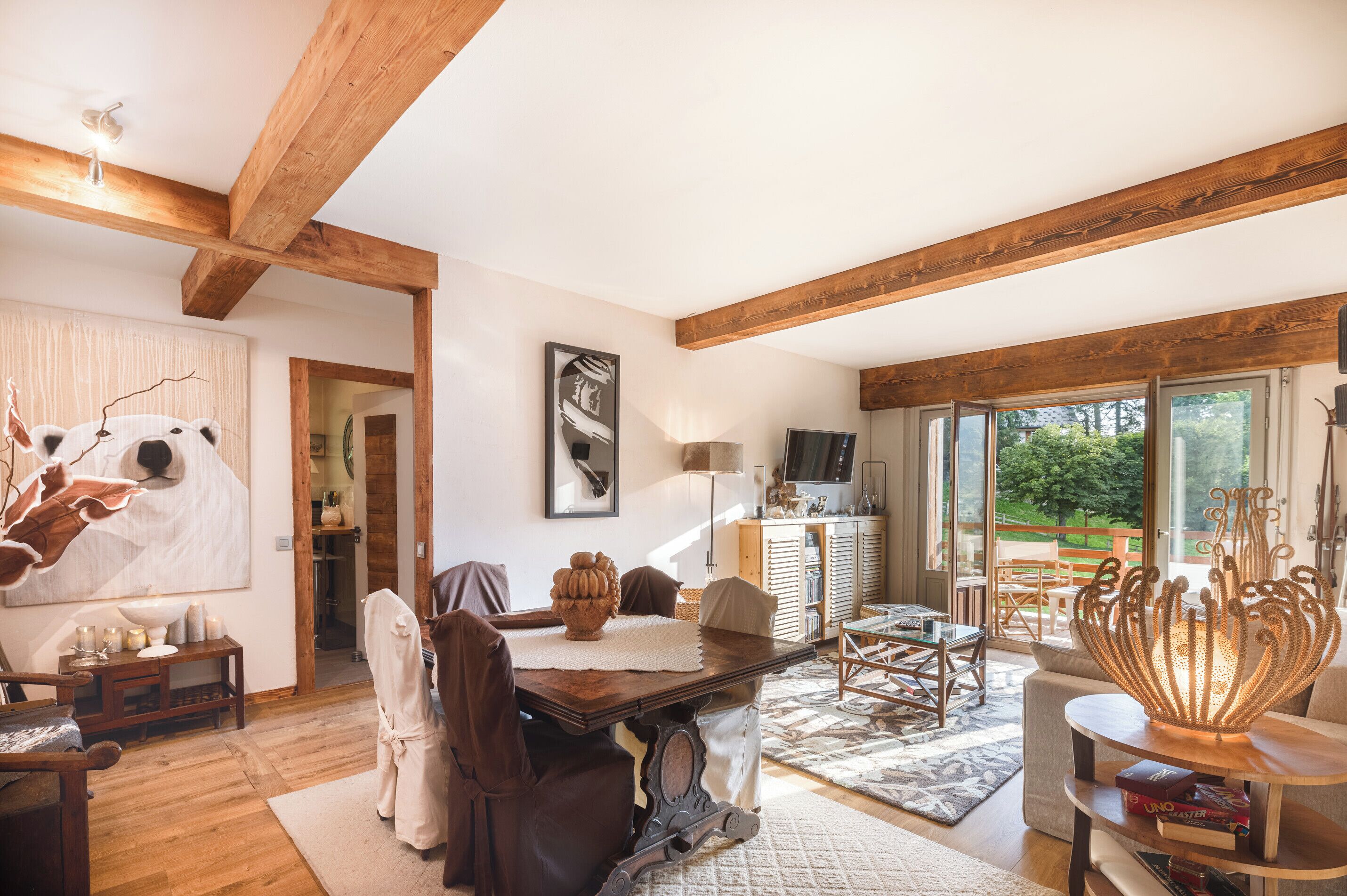 A spacious living room with wooden beams, a dining area, and a large window offering a view of the outdoors.