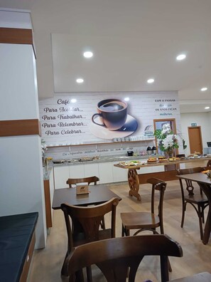 A café area with a large wall advertisement, wooden tables and chairs, and a counter with food items. - Hotel Maestro Executive Pato Branco (Pato Branco)