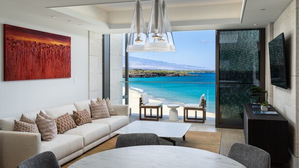 Un salon moderne doté d’une grande fenêtre donnant sur une plage, d’un canapé agrémenté de coussins à motifs, d’une table à manger et d’un téléviseur à écran plat fixé au mur.