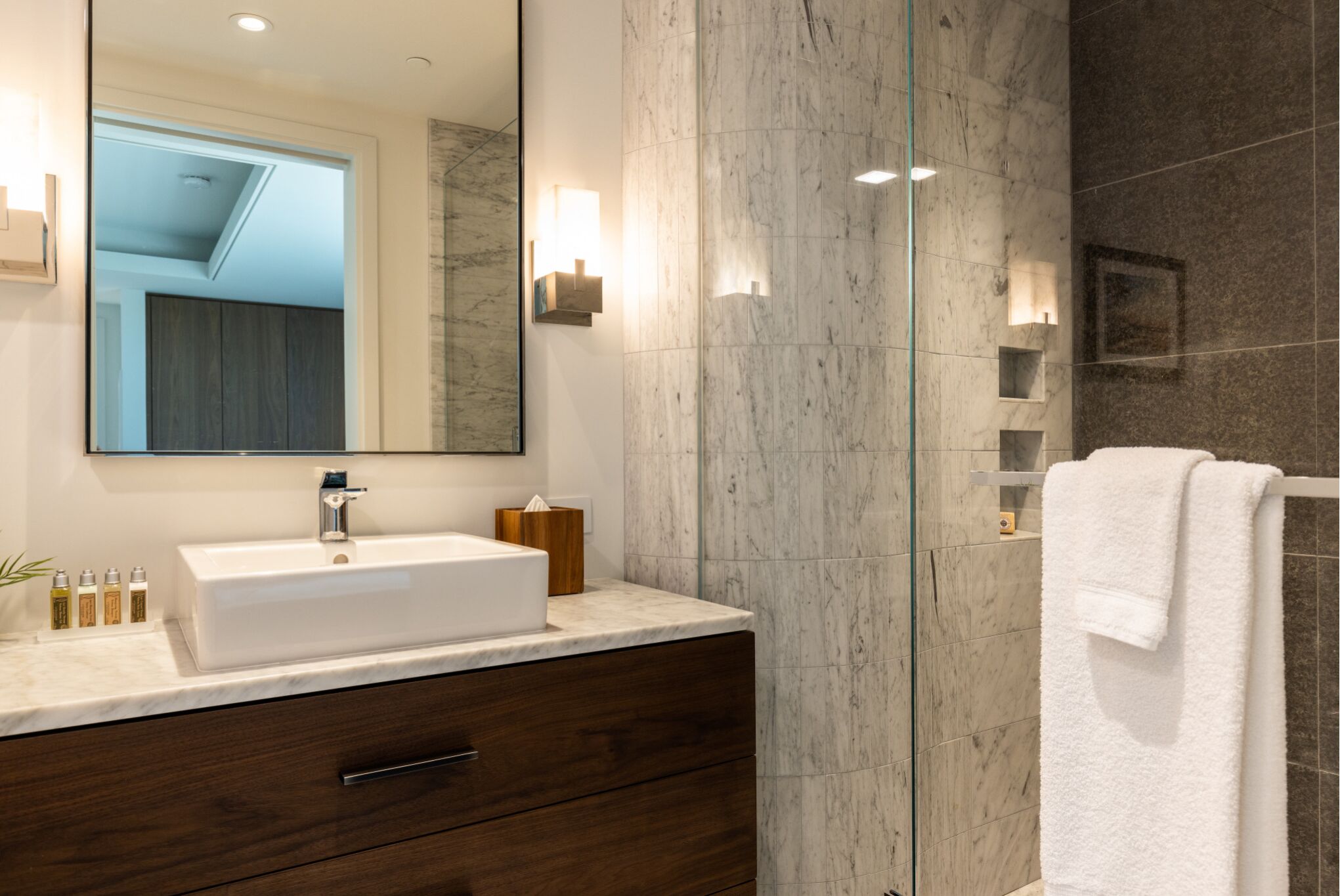 A modern bathroom with a large mirror, white sink, marble walls, and a towel rack.