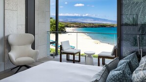Une chambre d’hôtel moderne dotée d’une grande fenêtre donnant sur une plage, d’un lit, d’une chaise et d’une petite table.