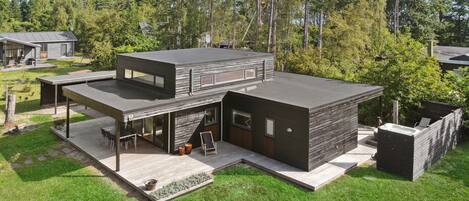 Una casa moderna de dos pisos con techo plano, ventanas grandes y una terraza con mobiliario exterior.