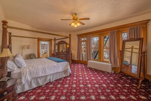 A bedroom with a four-poster bed, a radiator, a ceiling fan, and large windows with curtains.