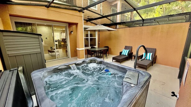 Una bañera de hidromasaje en un espacio exterior cubierto, con una mesa y sillas, y un ventanal corredizo que da a un espacio interior.