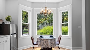 A dining area with a round table, chairs, and a chandelier. There are two windows with a view of greenery outside.