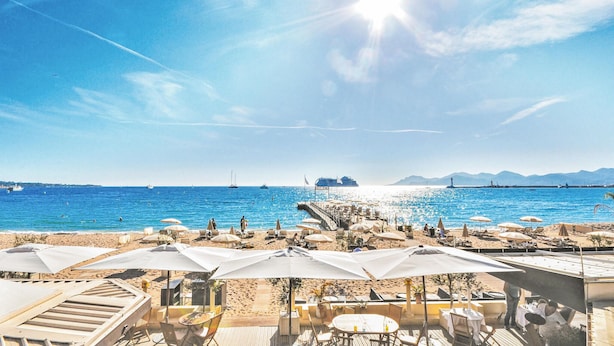 Un restaurant situé en bord de mer, avec une terrasse offrant une vue sur l’océan, une jetée et des bateaux en arrière-plan.