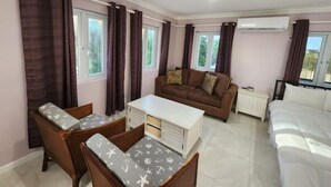 A room with a brown sofa, two wooden chairs, a white table, a bed, and large windows with curtains. - Periwinkle - Upscale Studio w Pool Near Beach
 (Nassau)