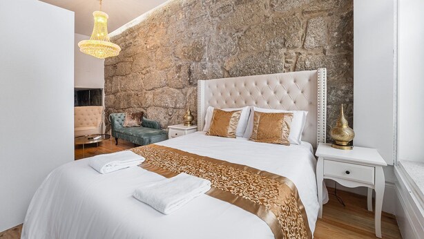 A bedroom with a stone wall, a tufted headboard, a large bed with white linens, a blue armchair, and a decorative lamp. - GuestReady - Boutique style loft in Constituição (Porto)