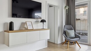 A modern living room with a large flat-screen TV mounted on the wall, a minimalist console table with a vase and a framed picture, a stylish grey armchair with a cushion, and a view of the outside through a large window with curtains. - GebirgsResort Winterberg (Winterberg)