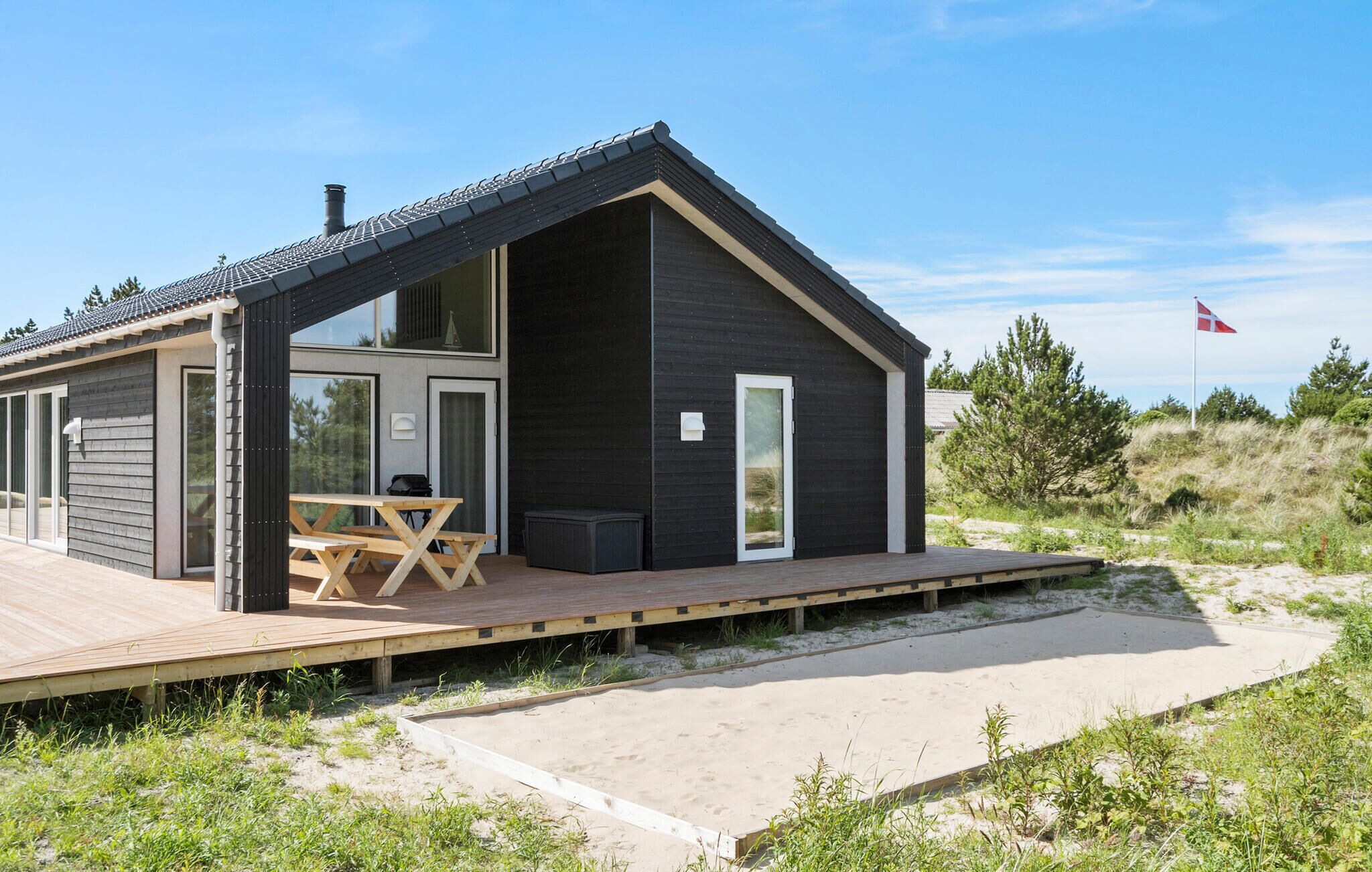 Et moderne sommerhus ved stranden med træterrasse, spiseområde og stort vindue.