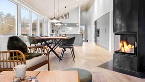 A modern kitchen with a large island, a dining area with a wooden table and chairs, and a fireplace with a visible fire. - Lovely home in Ringkøbing with kitchen (Ringkøbing)