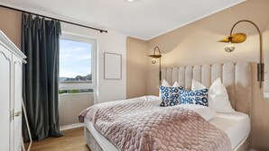 A bedroom with a large bed, a headboard with multiple pillows, a bedside lamp, and a view through a window with curtains. - Gorgeous apartment in Winterberg (Winterberg)