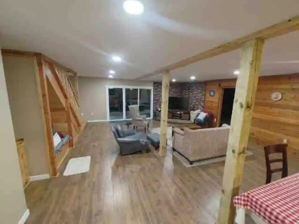 Un vaste espace de vie doté d’un plancher en bois, d’un canapé, d’une table à manger recouverte d’une nappe à carreaux, et d’un coin cuisine avec des murs en bois et un pare-feu en briques.